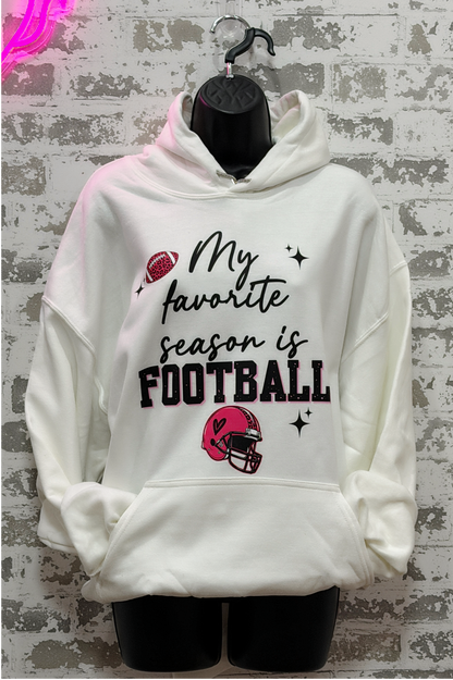 A white pullover hoodie displayed on a black mannequin torso against a distressed gray brick wall. The hoodie features a graphic print that reads 'My favorite season is FOOTBALL' in black script and block lettering, accompanied by illustrations of a red football and a red football helmet.