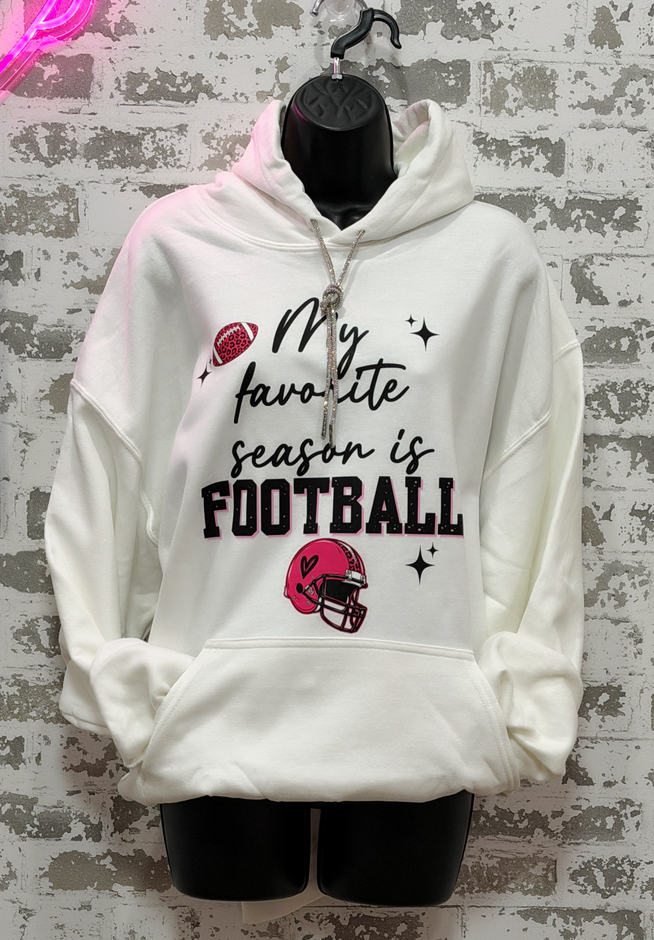 A white pullover hoodie displayed on a black mannequin torso against a distressed gray brick wall. The hoodie features a graphic print that reads 'My favorite season is FOOTBALL' in black script and block lettering, accompanied by a red football, a red helmet, and small black star accents.