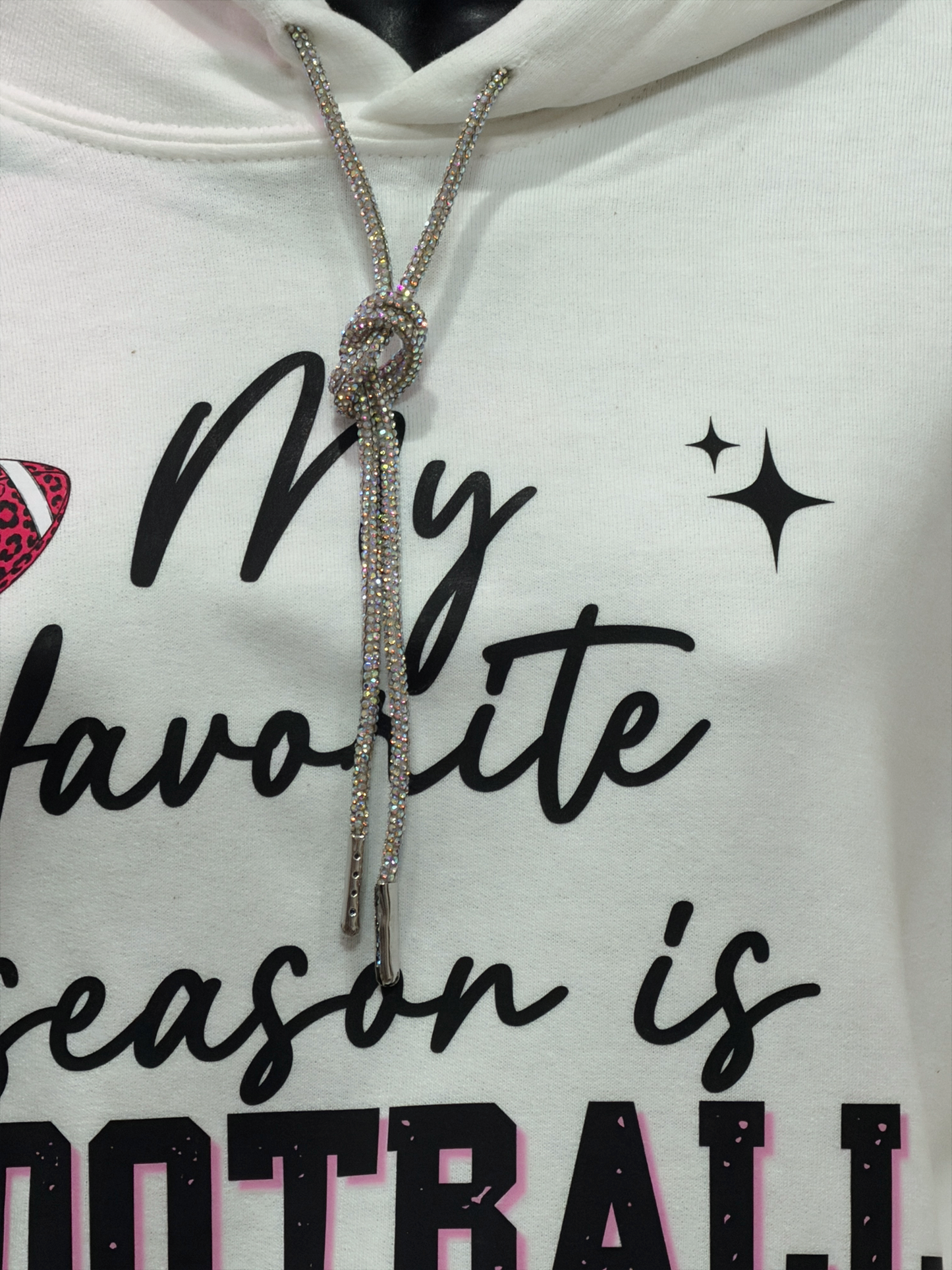 A close-up of a white hoodie featuring the text 'My favorite season is FOOTBALL' in black and pink lettering, with a sparkling silver rhinestone drawstring knot at the collar.