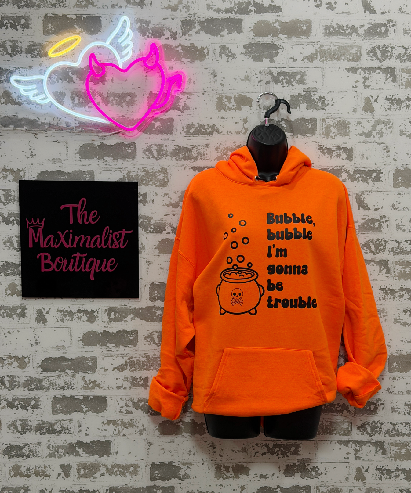 Bright orange pullover hoodie on a black hanger against a distressed white brick wall, featuring a black graphic of a cauldron and the text 'Bubble, bubble I'm gonna be trouble'. A neon angel sign and a black sign reading 'The Maximalist Boutique' are visible on the left.