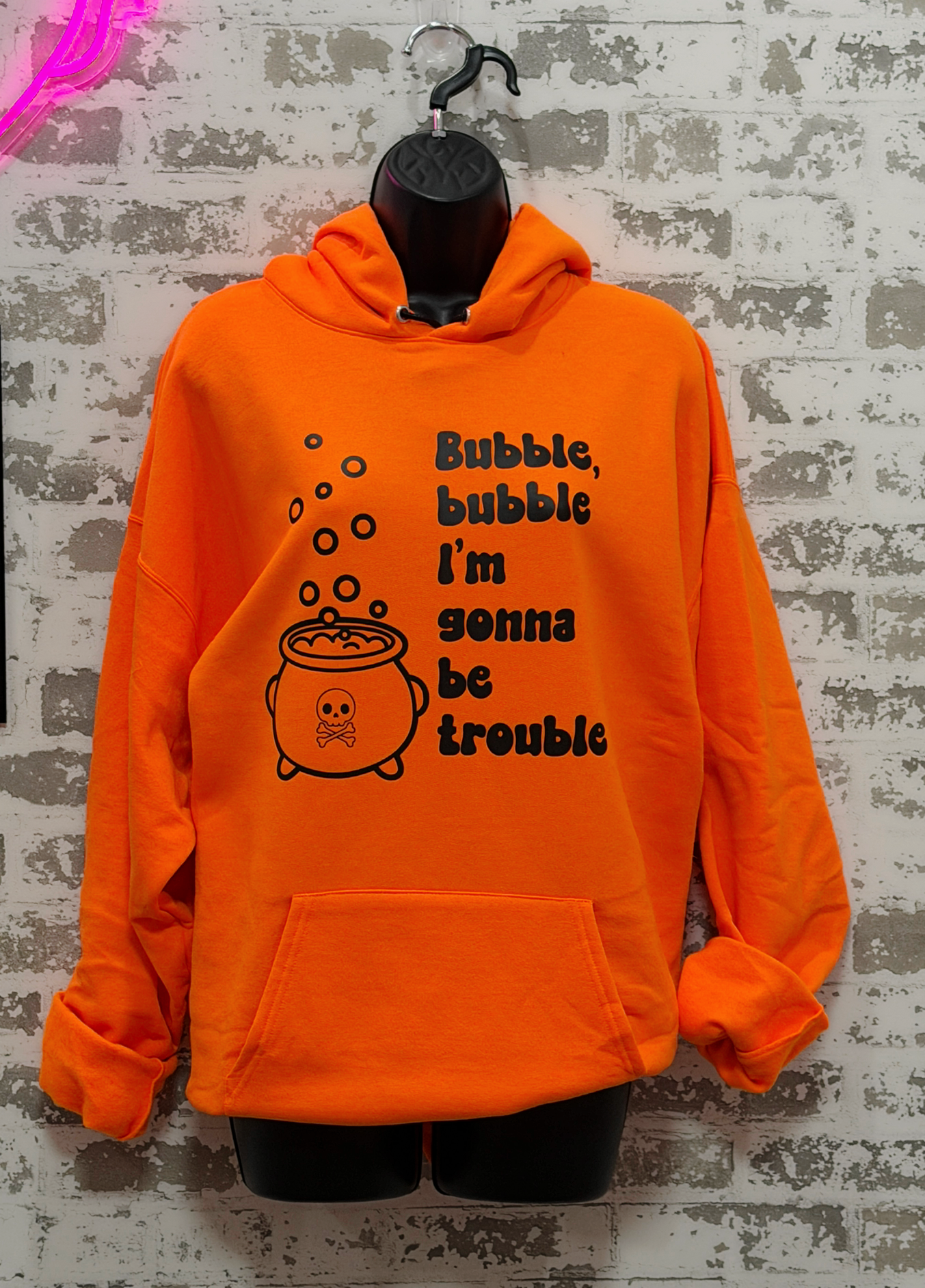 Bright orange pullover hoodie displayed on a black mannequin torso against a distressed white brick wall. The hoodie features a black graphic of a cauldron with bubbles and the text 'Bubble, bubble I'm gonna be trouble'.