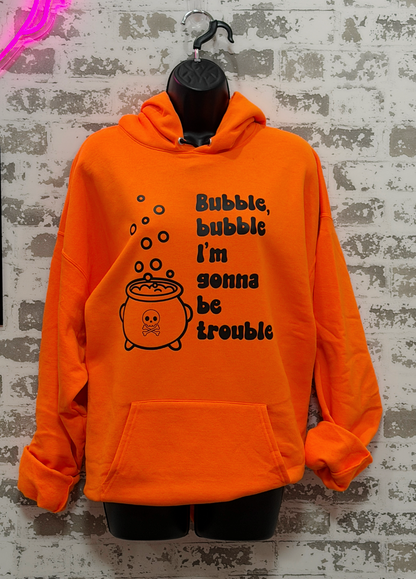 Bright orange pullover hoodie displayed on a black mannequin torso against a distressed white brick wall. The hoodie features a black graphic of a cauldron with bubbles and the text 'Bubble, bubble I'm gonna be trouble'.