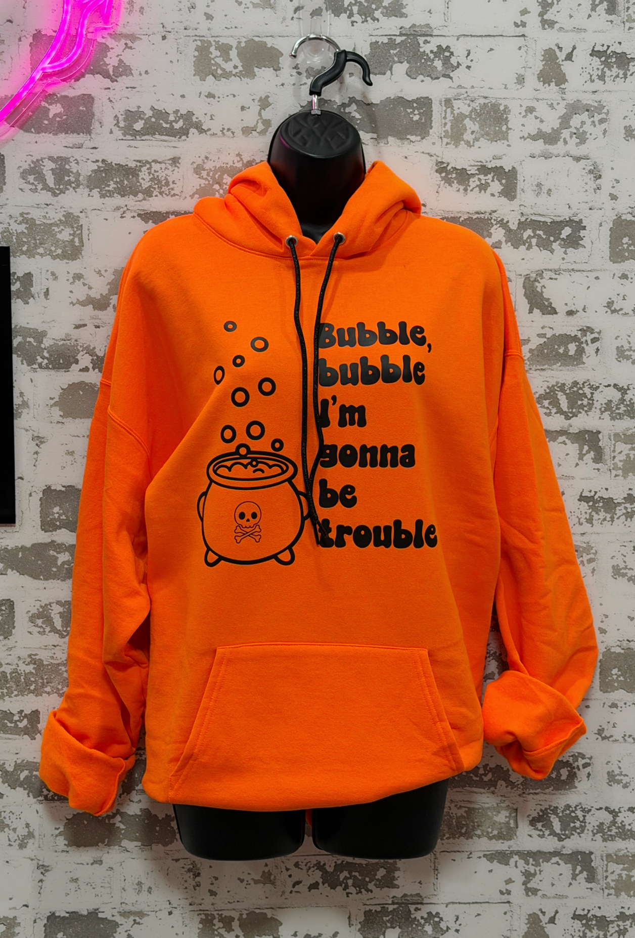 Bright orange pullover hoodie on a black hanger and mannequin torso, featuring a black graphic of a cauldron with bubbles and the text 'Bubble, bubble I'm gonna be trouble' on the front, set against a distressed white brick wall.
