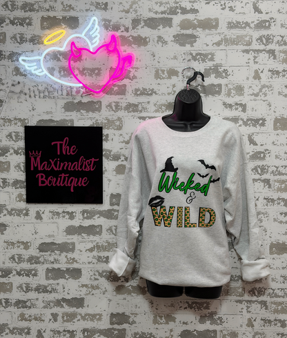 A light gray long-sleeve crewneck sweatshirt with a 'Wicked WILD' graphic on a black hanger, displayed against a faux brick wall with a neon angel sign and a boutique logo.