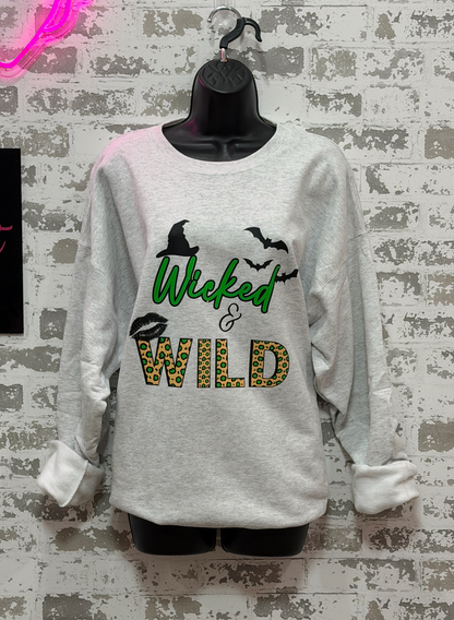 A light heather gray long-sleeve t-shirt with a 'Wicked & Wild' graphic print, displayed on a black mannequin torso against a distressed white brick wall background.