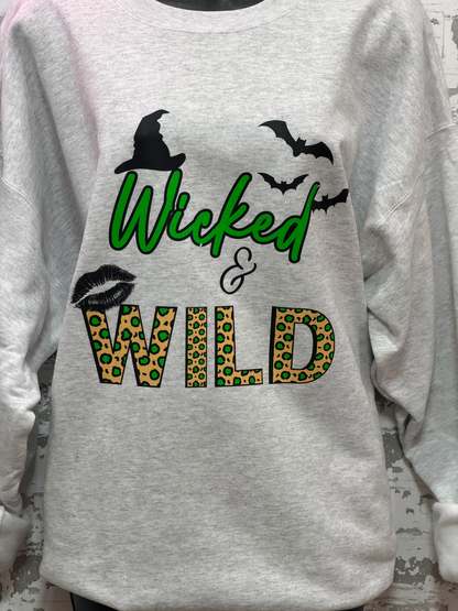 A light heather gray long-sleeve sweatshirt featuring a Halloween-themed graphic print with the words 'Wicked & Wild' in green and leopard-print lettering, decorated with black witch hats, bats, lips, and an ampersand.