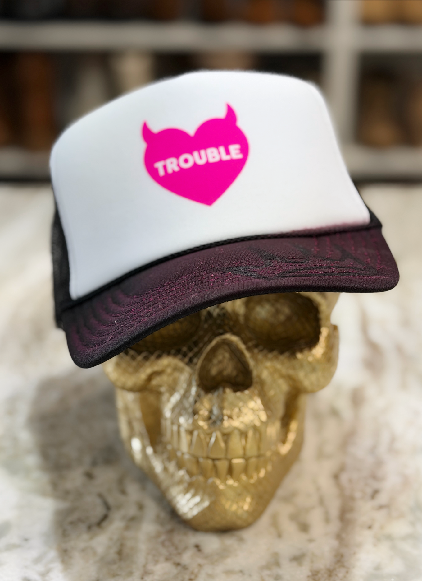 A white and dark burgundy trucker hat with a hot pink devil-heart logo that says 'TROUBLE' in white, displayed on a gold skull.