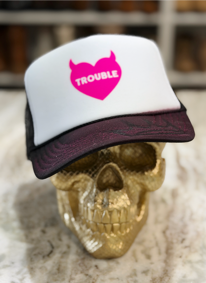 A white and dark burgundy trucker hat with a hot pink devil-heart logo that says 'TROUBLE' in white, displayed on a gold skull.