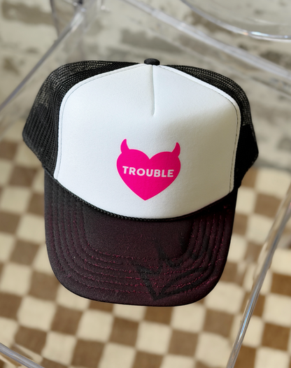 A black and white trucker hat with a bright pink heart graphic on the front that reads 'TROUBLE' in white text, displayed on a clear acrylic chair over a checkered floor.