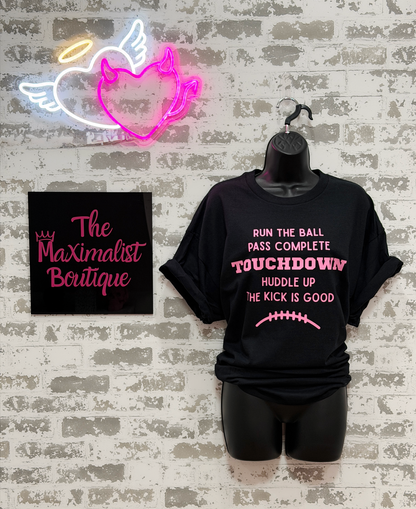 A black short-sleeve graphic T-shirt displayed on a black mannequin torso against a faux white brick wall. The shirt has pink and white text reading 'RUN THE BALL PASS COMPLETE TOUCHDOWN HUDDLE UP THE KICK IS GOOD' with a small football graphic below.
