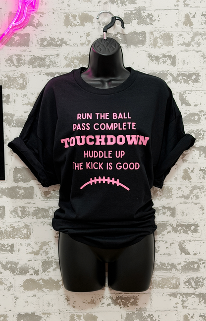A black short-sleeve graphic T-shirt displayed on a black mannequin torso against a distressed white brick wall. The shirt has pink text reading 'RUN THE BALL PASS COMPLETE TOUCHDOWN Huddle Up The Kick Is Good' with a pink football graphic below.
