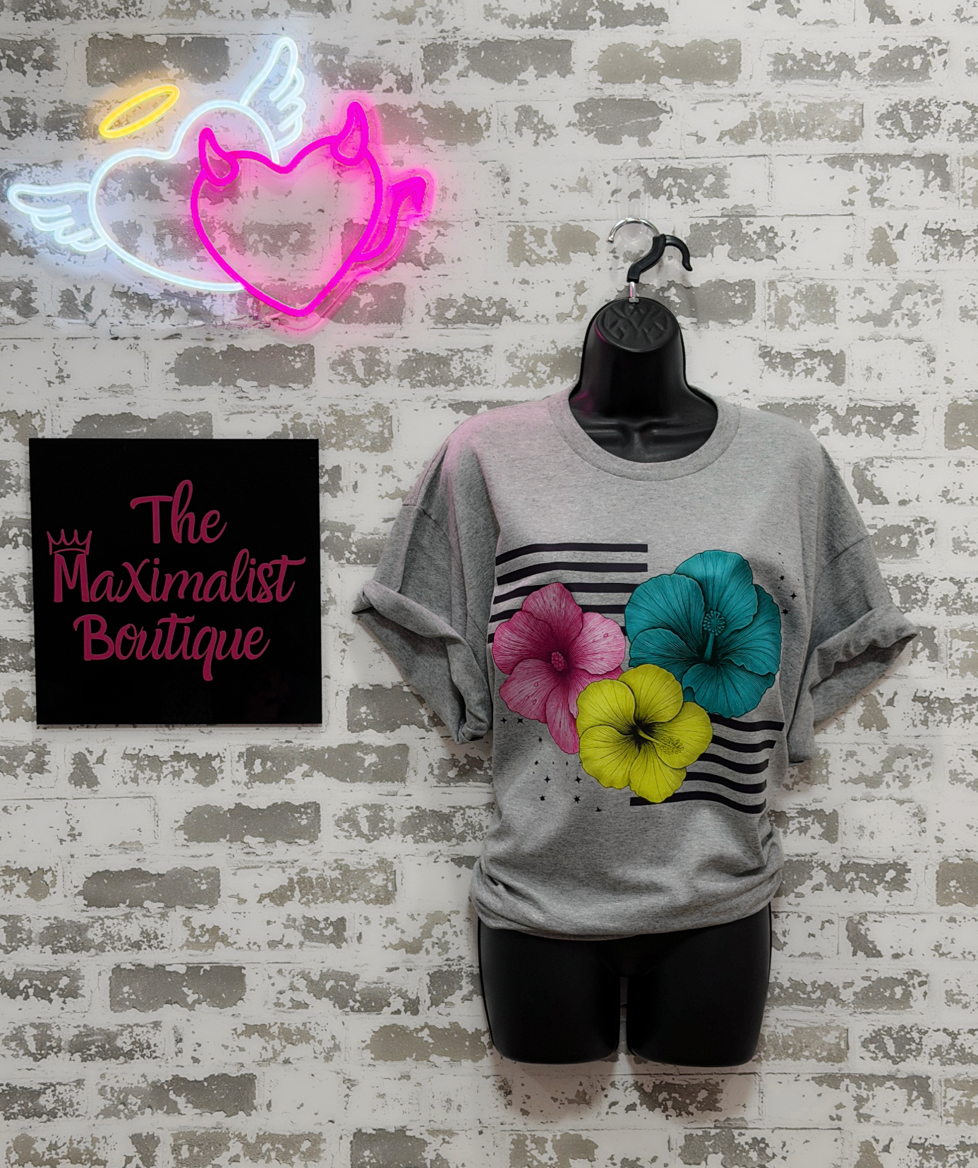 A gray short-sleeve t-shirt displayed on a black mannequin torso against a white brick wall. The shirt features a graphic print with black horizontal stripes, a pink hibiscus flower, a teal flower, and a yellow flower.