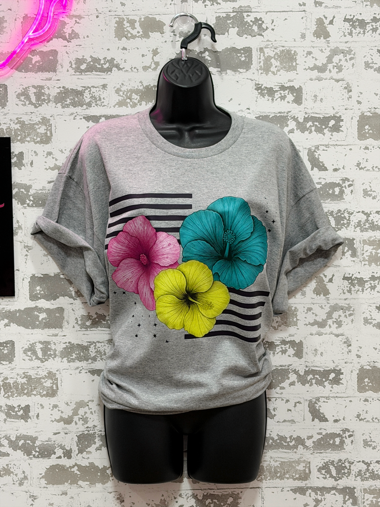 A heather gray short-sleeve t-shirt displayed on a black mannequin torso against a distressed white brick wall. The shirt features a graphic print of three hibiscus flowers in pink, teal, and yellow over horizontal black stripes.