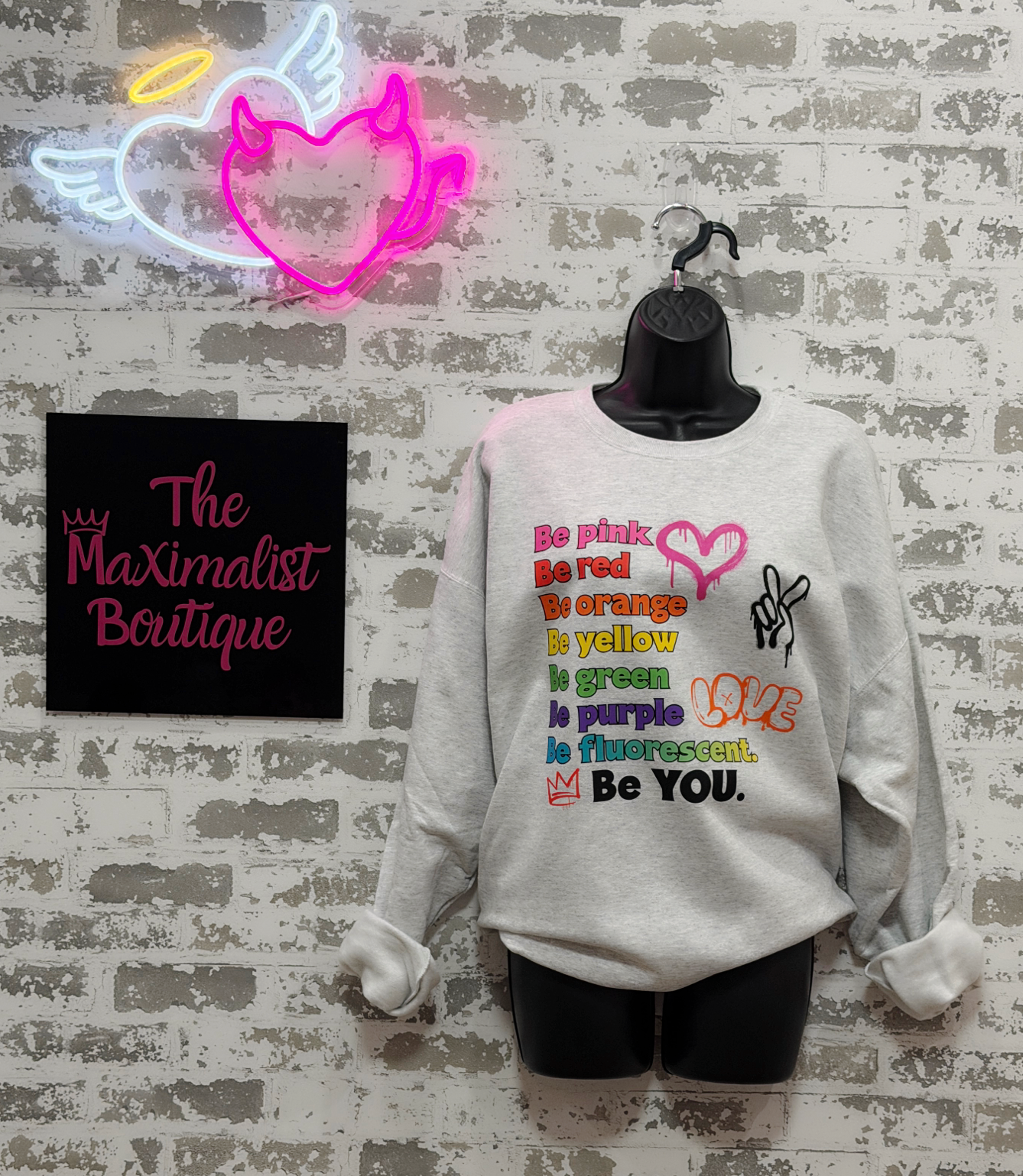A light gray crewneck sweatshirt displayed on a black mannequin torso against a faux brick wall. The sweatshirt features a colorful retro-style graphic with text and rainbow-colored symbols.