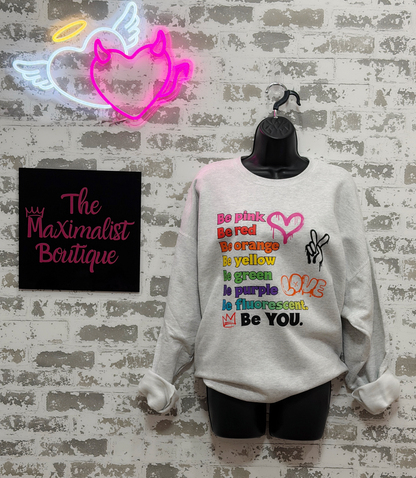 A light gray crewneck sweatshirt displayed on a black mannequin torso against a faux brick wall. The sweatshirt features a colorful retro-style graphic with text and rainbow-colored symbols.