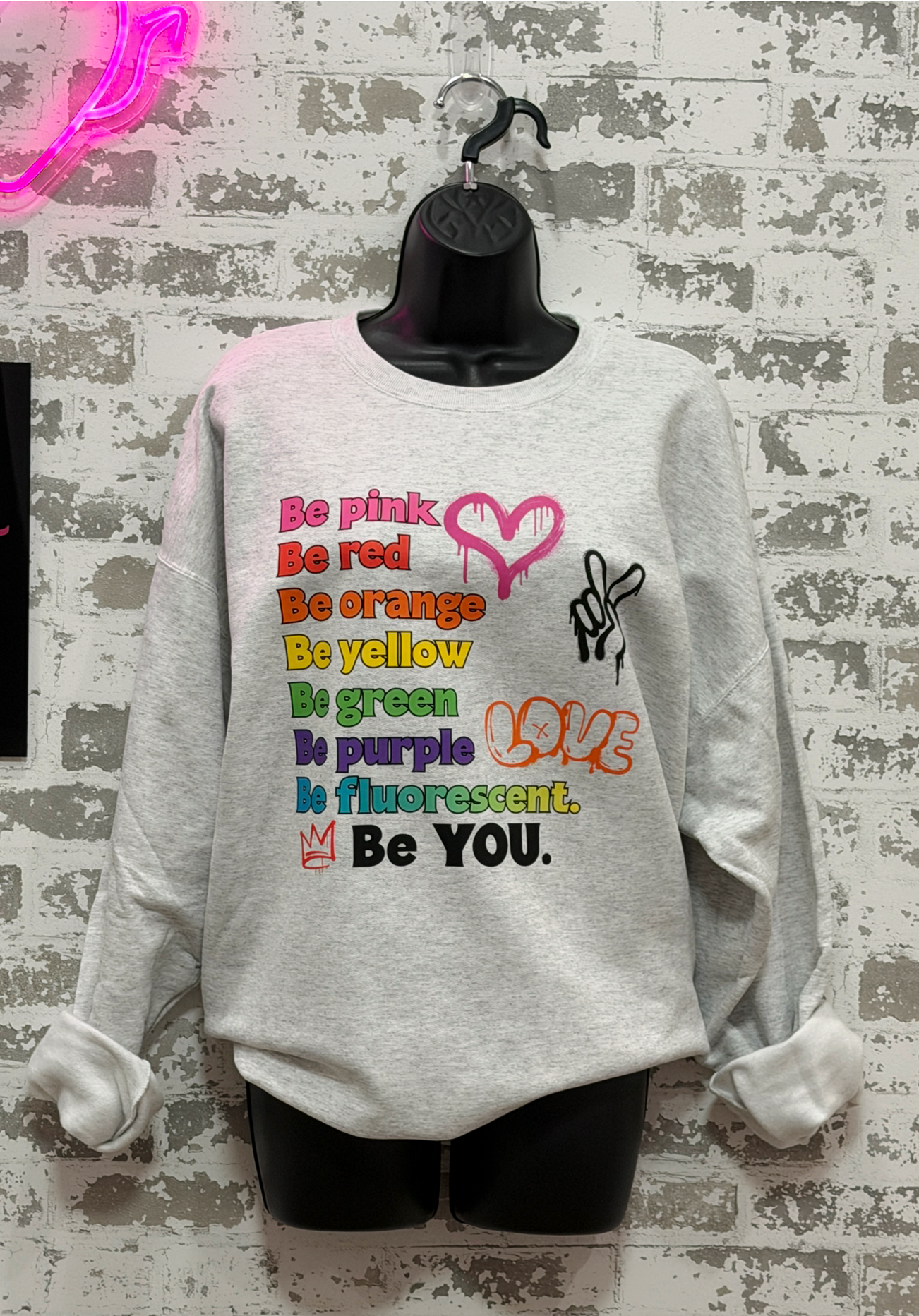A light heather gray sweatshirt displayed on a black mannequin torso against a distressed white brick wall. The sweatshirt features a colorful graphic text print reading 'Be pink Be red Be orange Be yellow Be green Be blue Be purple Be fluorescent.