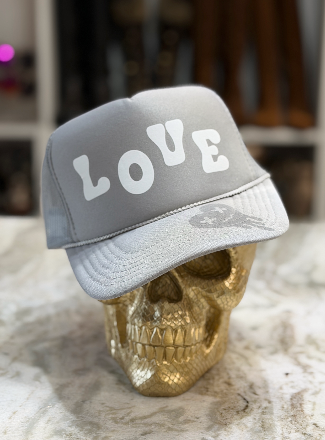 A light gray trucker-style cap with the word 'LOVE' in large white letters on the front, displayed on a metallic gold skull against a softly blurred indoor background.