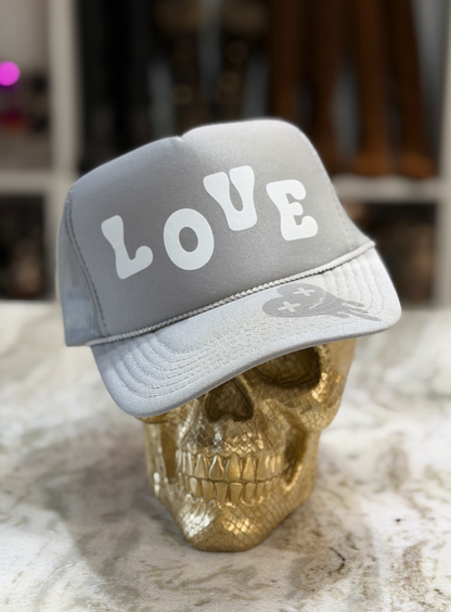 A light gray trucker-style cap with the word 'LOVE' in large white letters on the front, displayed on a metallic gold skull against a softly blurred indoor background.