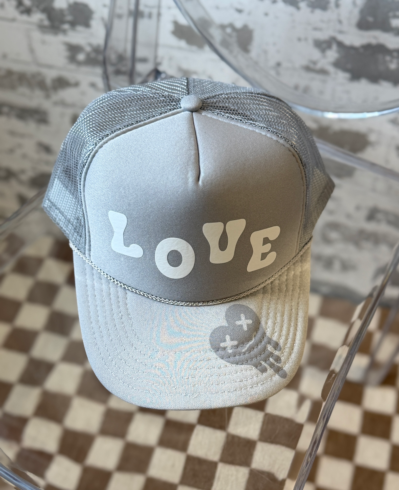 A light gray trucker cap with a gray mesh back, featuring the word 'LOVE' in white letters across the front and a gray tired heart graphic on the brim, displayed on a clear acrylic stand over a checkered floor.