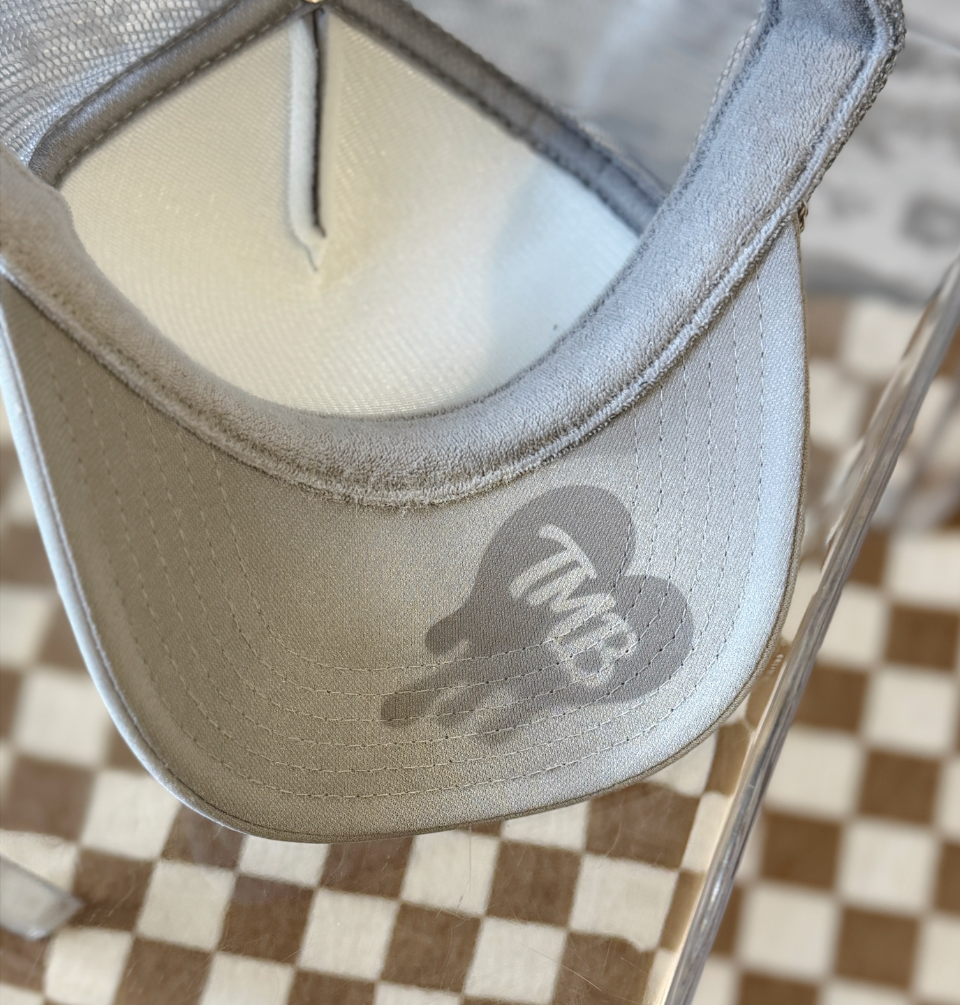 Close-up of the inside of a light gray trucker-style cap with a gray TMB logo printed on the brim, resting on a clear acrylic stand over a brown-and-white checkered surface.