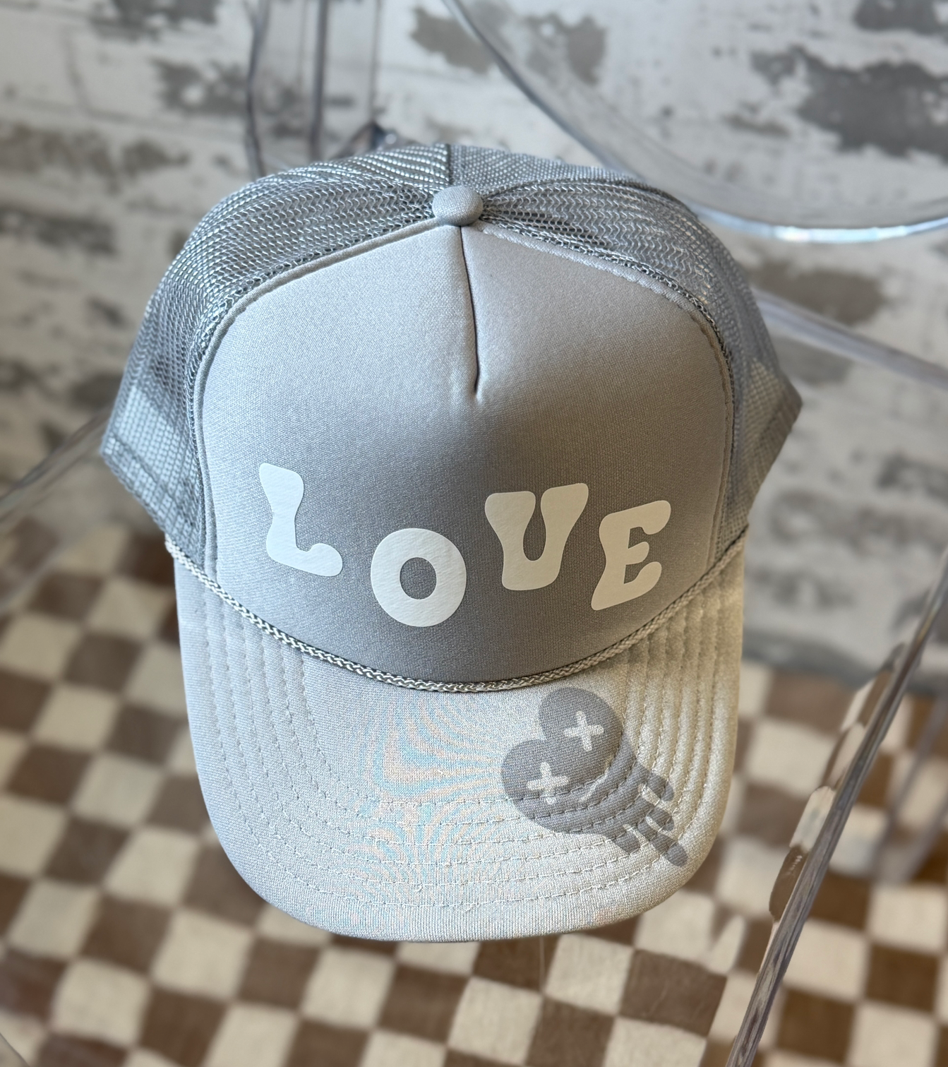 A light gray trucker hat with a mesh back, featuring the word 'LOVE' in white letters across the front panels and a small gray tired heart graphic on the brim.