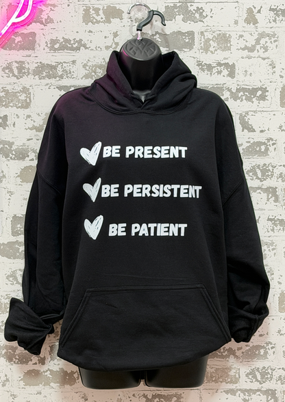 A black pullover hoodie displayed on a black hanger/mannequin against a faux brick wall background. The hoodie features three white printed phrases with heart icons: 'BE PRESENT', 'BE PERSISTENT', and 'BE PATIENT'.