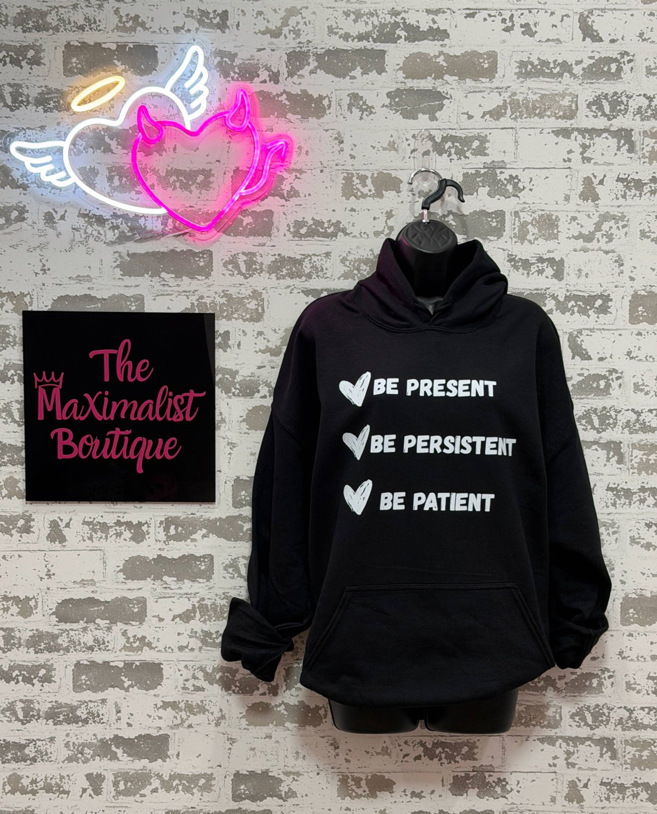 A black pullover hoodie hanging on a black hanger against a distressed white brick wall. The hoodie has white text listing 'BE PRESENT', 'BE PERSISTENT', and 'BE PATIENT' with white heart icons.