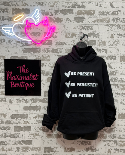 A black pullover hoodie hanging on a black hanger against a distressed white brick wall. The hoodie has white text listing 'BE PRESENT', 'BE PERSISTENT', and 'BE PATIENT' with white heart icons.