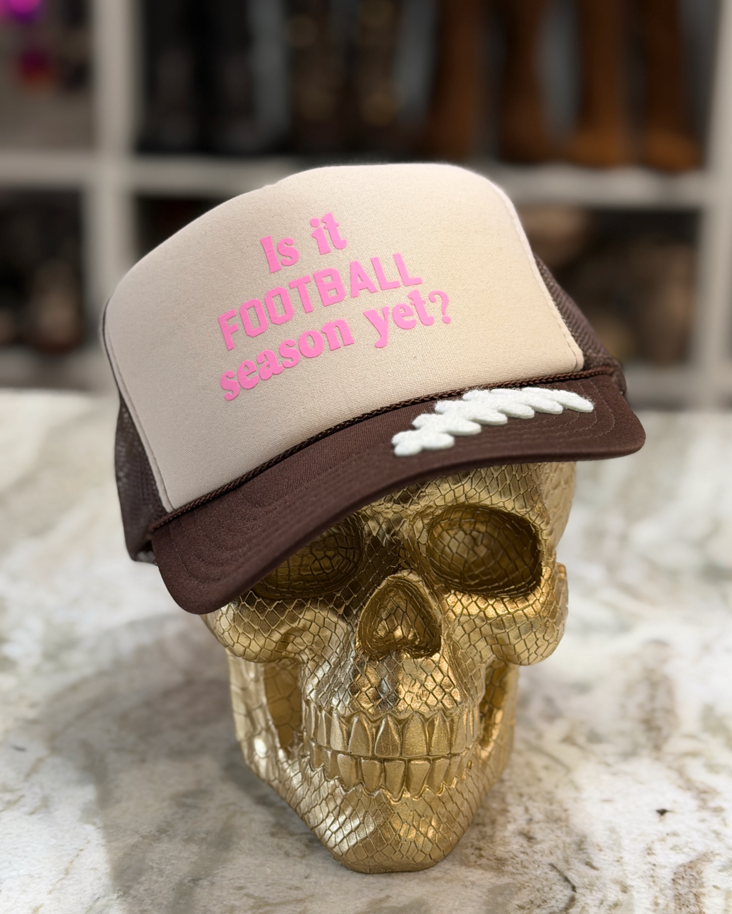 A beige and brown trucker hat with pink embroidered text reading 'Is it FOOTBALL season yet?' and white football-shaped accents on the brim, displayed on a gold skull mannequin against a blurred indoor background.
