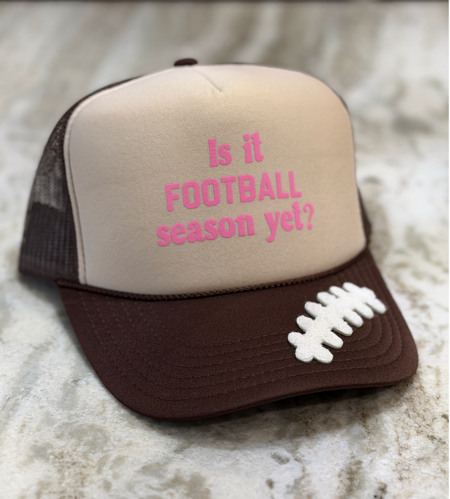 A trucker-style baseball cap with a beige front panel, brown mesh back, and brown visor. The front panel has pink embroidered text reading 'Is it FOOTBALL season yet?' and a white football laces graphic on the brim.
