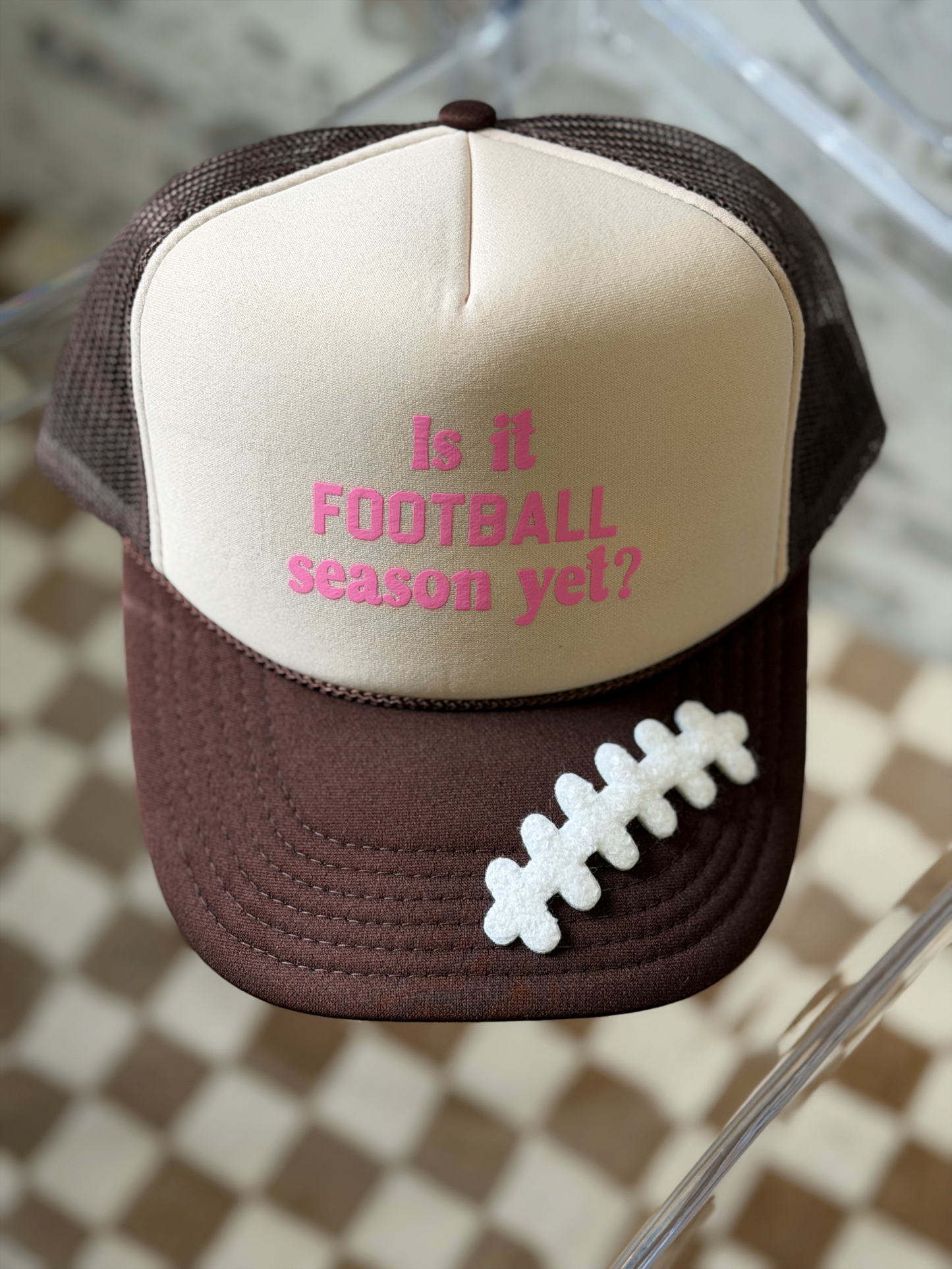 A brown and beige trucker hat with pink embroidered text reading 'Is it FOOTBALL season yet?' and a white football patch on the brim, displayed on a glass surface over a checkered floor.