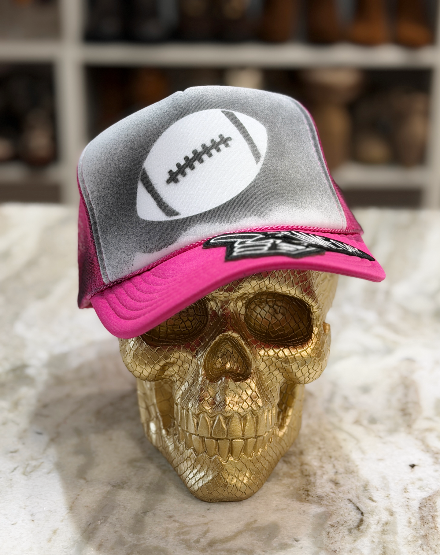 A white and pink trucker-style football cap with a white football graphic on the front, displayed on a metallic gold skull against a blurred indoor background.