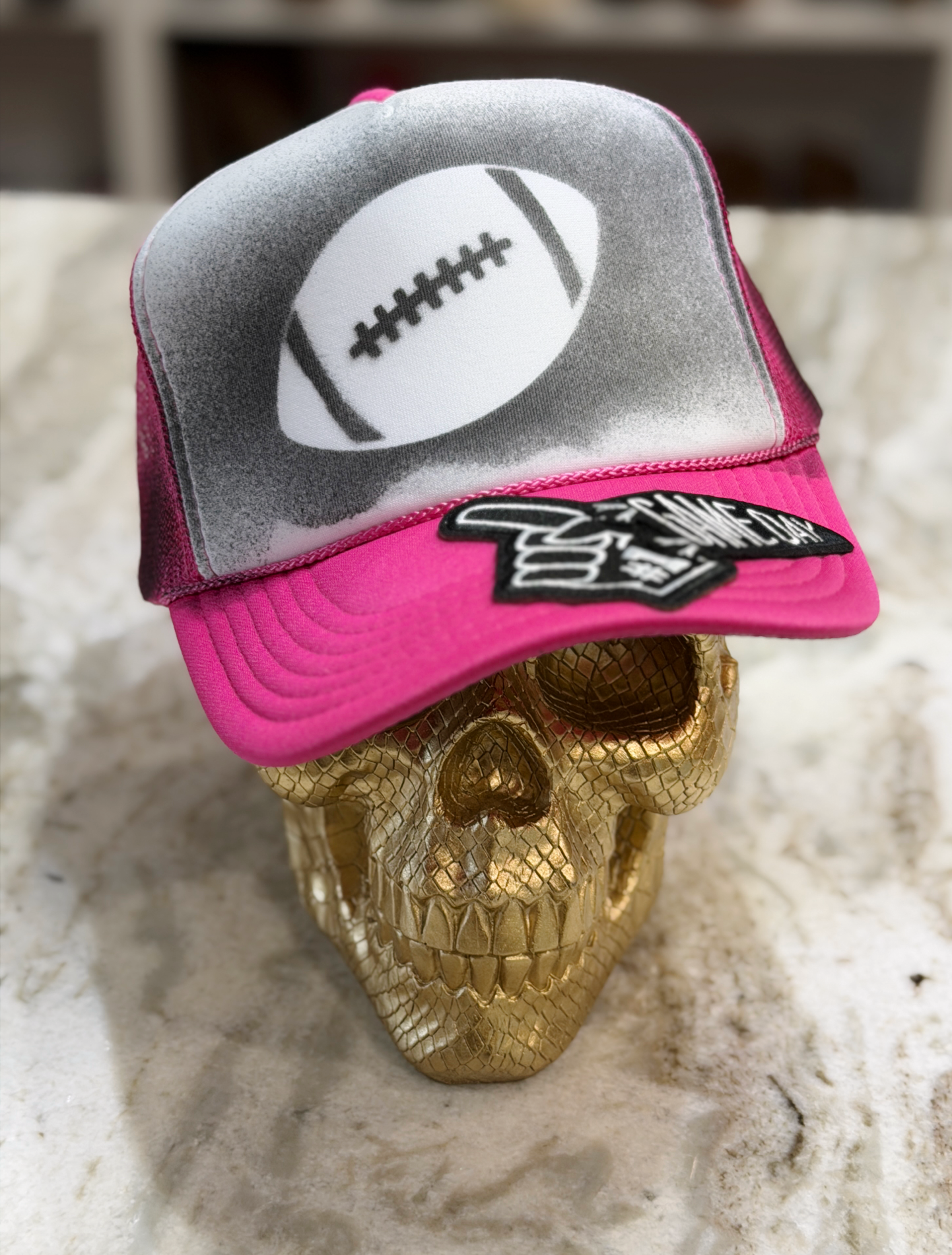 A pink and gray trucker-style cap with a football graphic on the front and a black-and-white patch on the brim, displayed on a gold skull against a light stone surface.