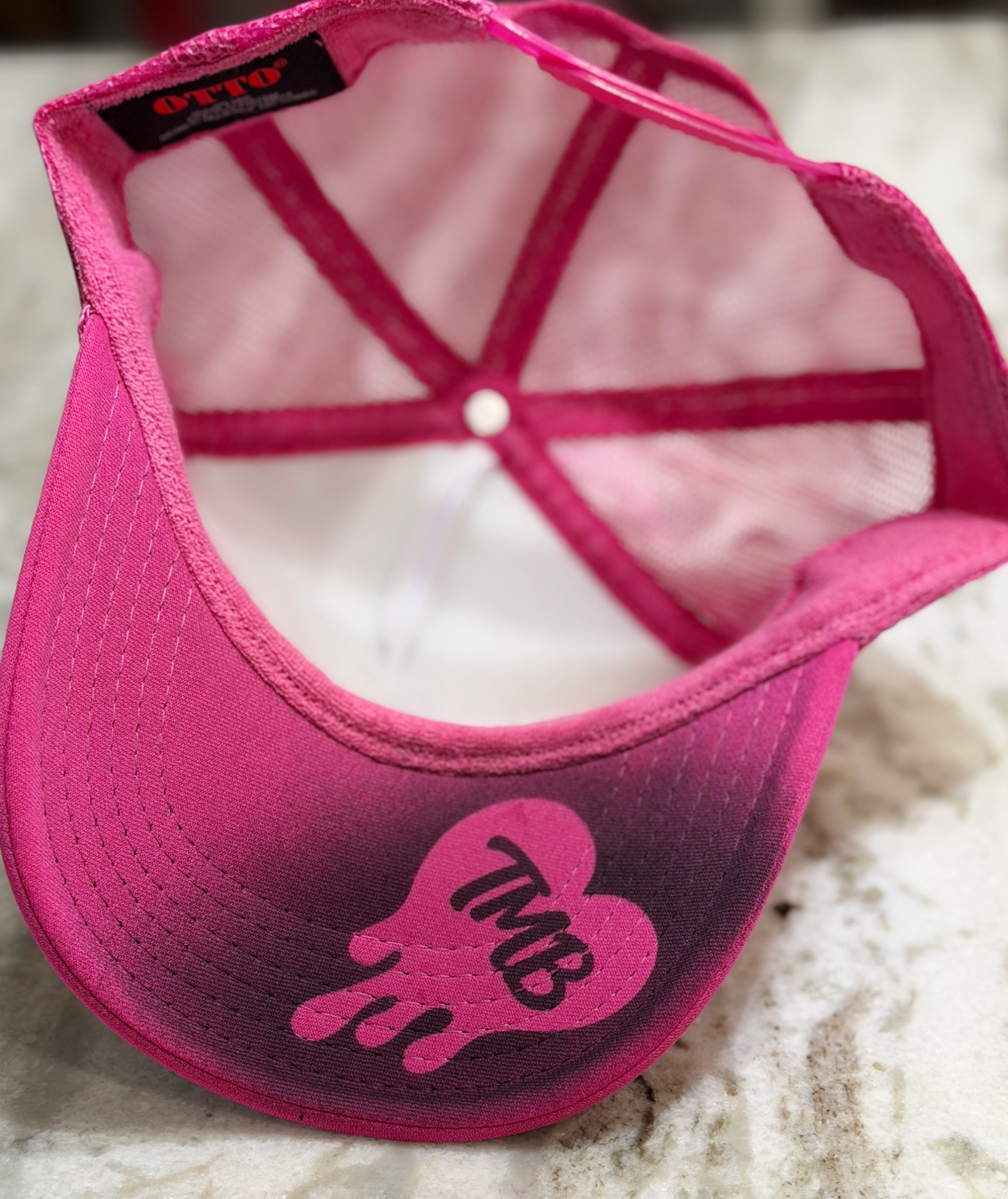 A pink snapback cap with a gradient fade from bright pink to dark purple, featuring a pink heart-shaped graphic with the letters 'TMB' on the brim. The inside of the cap shows a pink mesh back and a black brand label reading 'OTTO'.