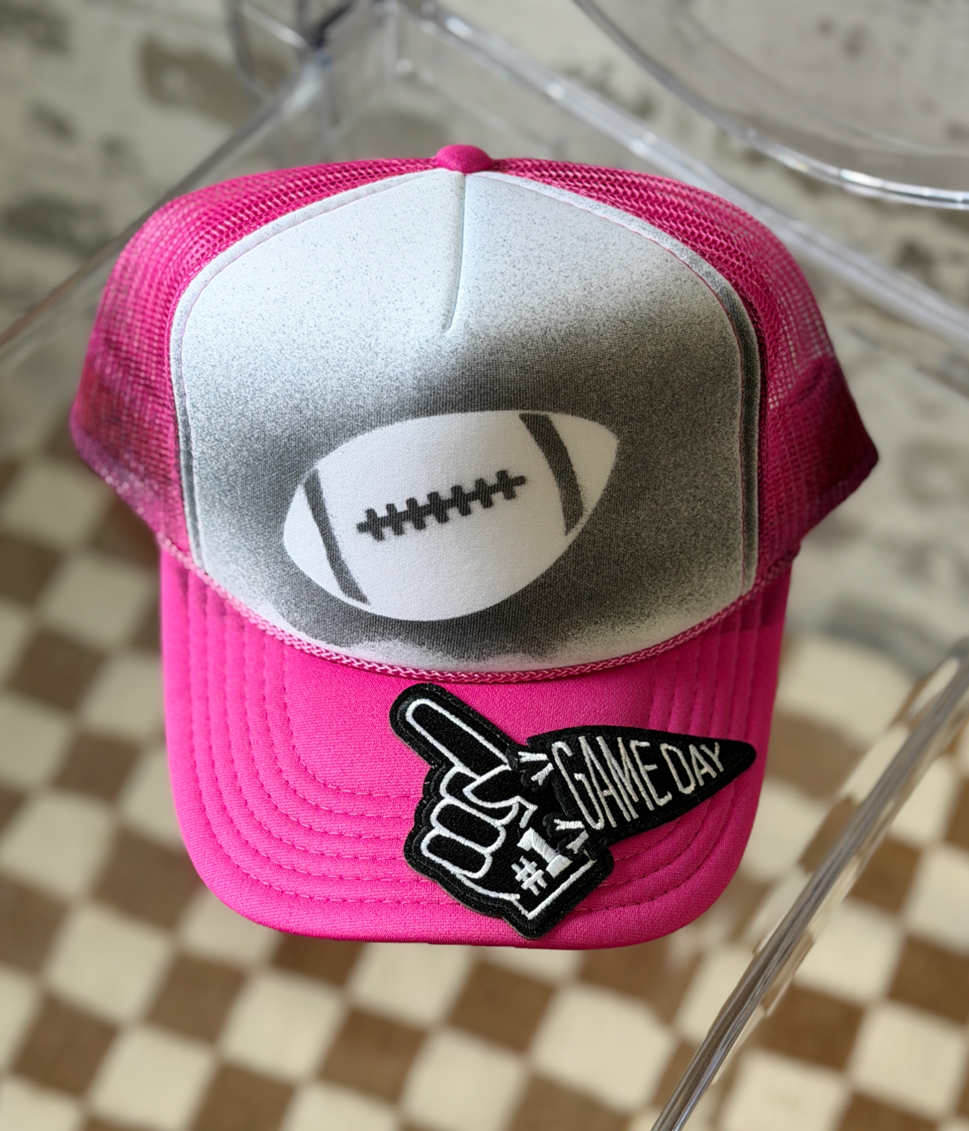 A pink and silver trucker hat with a football graphic on the front panel and a black-and-white 'GAME DAY #1' foam finger patch on the brim, displayed on a clear chair over a checkered floor.