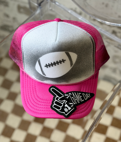 A pink and silver trucker hat with a football graphic on the front panel and a black-and-white 'GAME DAY #1' foam finger patch on the brim, displayed on a clear chair over a checkered floor.