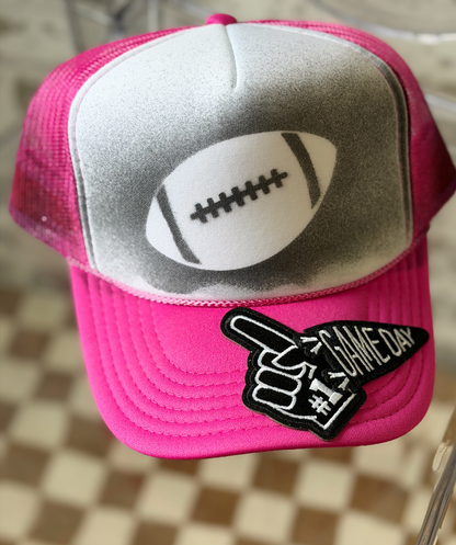 A pink and white trucker-style baseball cap featuring a football graphic on the front panel and a black-and-white 'GAME DAY' patch on the brim, photographed against a checkered floor background.