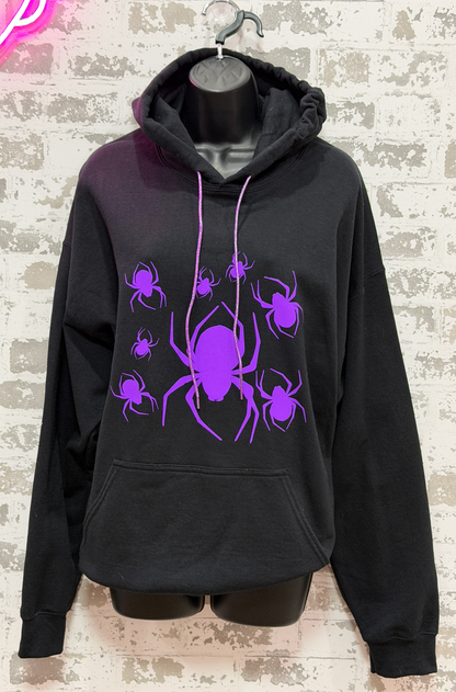 A black pullover hoodie displayed on a black mannequin torso against a light brick wall. The hoodie features a large purple spider graphic with smaller spiders surrounding it on the front, along with matching purple drawstrings.