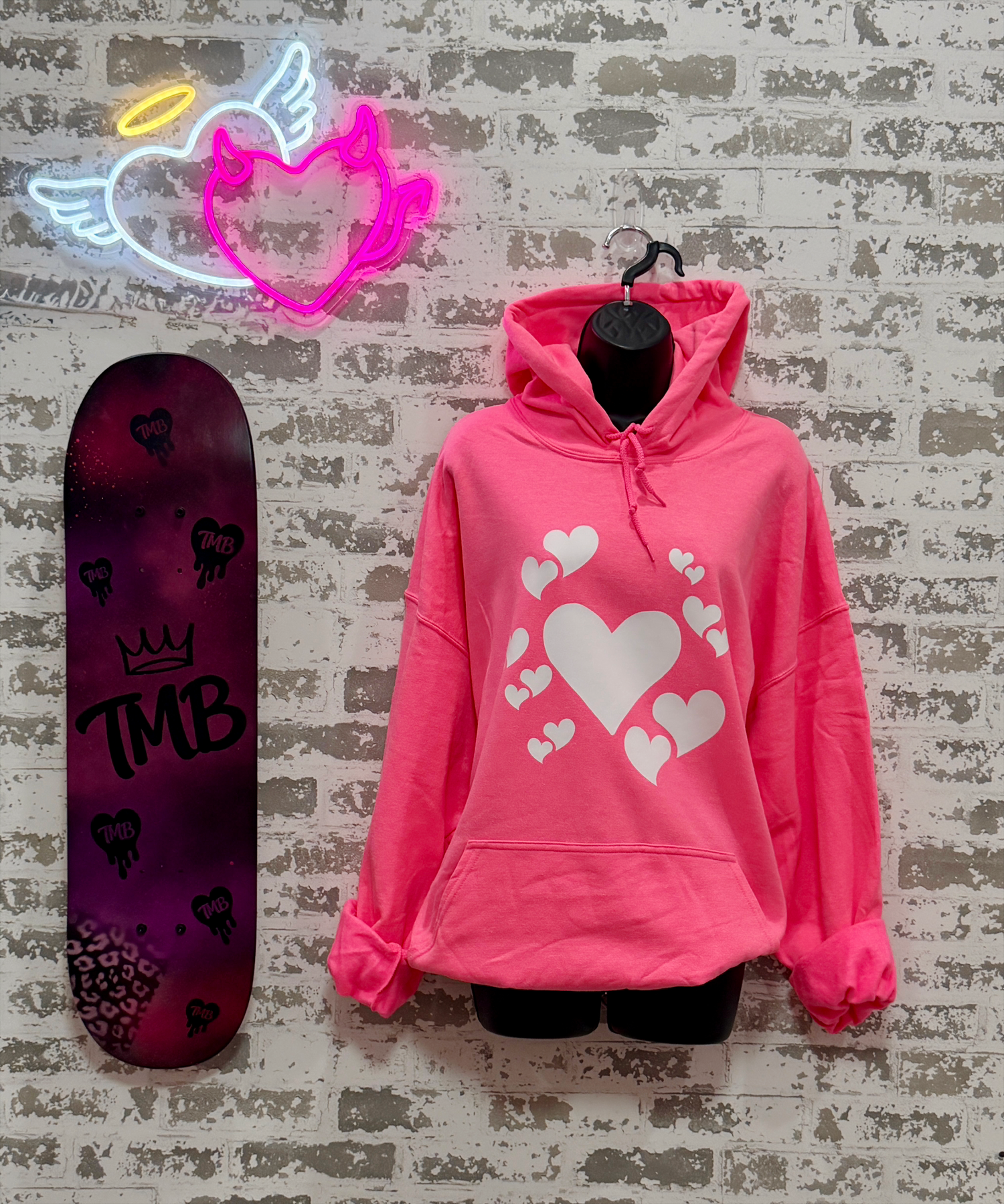 A bright pink pullover hoodie with a white heart graphic print, displayed on a black hanger against a distressed white brick wall. A neon angel heart sign glows above, and a purple skateboard with 'TMB' branding leans against the wall on the left.