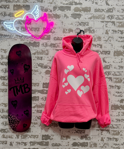 A bright pink pullover hoodie with a white heart graphic print, displayed on a black hanger against a distressed white brick wall. A neon angel heart sign glows above, and a purple skateboard with 'TMB' branding leans against the wall on the left.