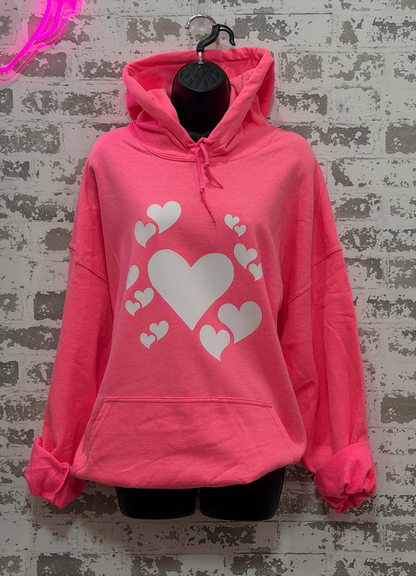 A bright pink pullover hoodie with a white heart graphic print on the front, displayed on a black mannequin torso against a distressed gray brick wall.