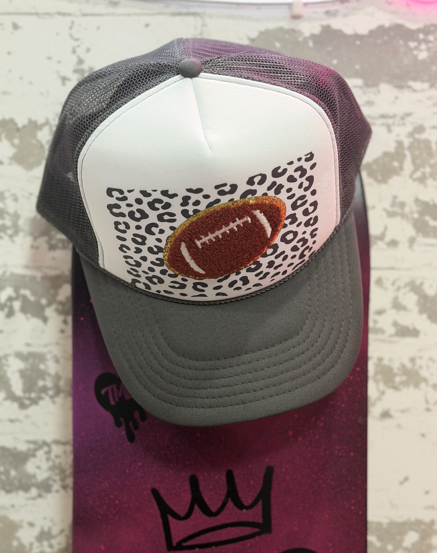 A gray and white trucker hat with a football patch on a leopard print background, displayed on a purple skateboard against a distressed white wall.