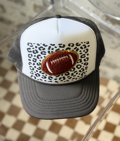 A trucker-style baseball cap with a white front panel, black mesh back, and a leopard print pattern on the front. The center features an embroidered brown football patch outlined in gold glitter.