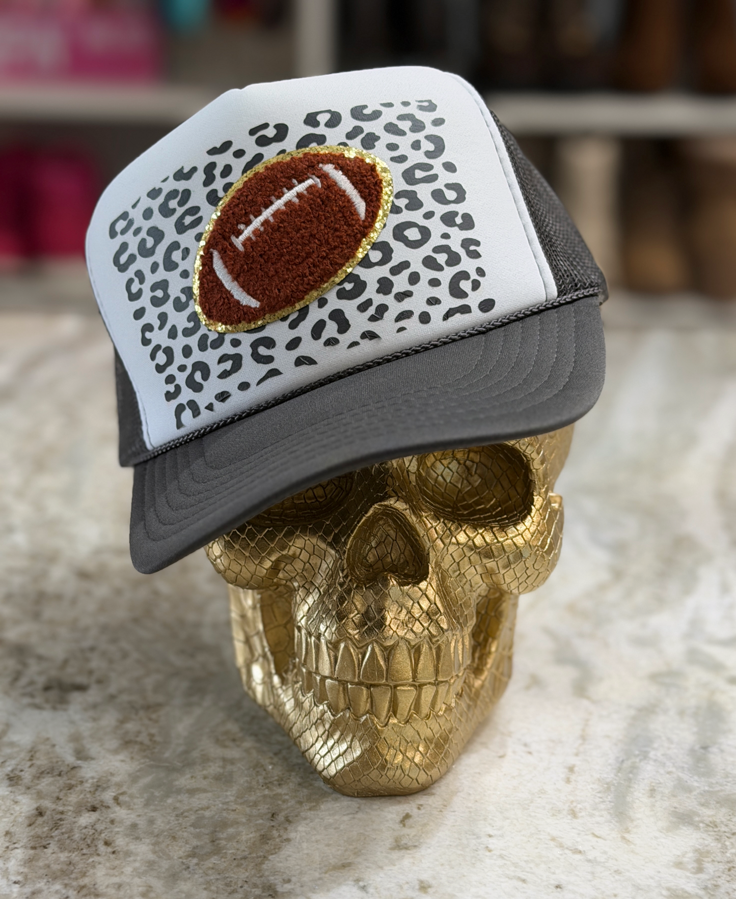 A trucker-style cap with a white front panel featuring a black leopard print pattern and a brown embroidered football patch outlined in gold, displayed on a metallic gold skull against a blurred indoor background.