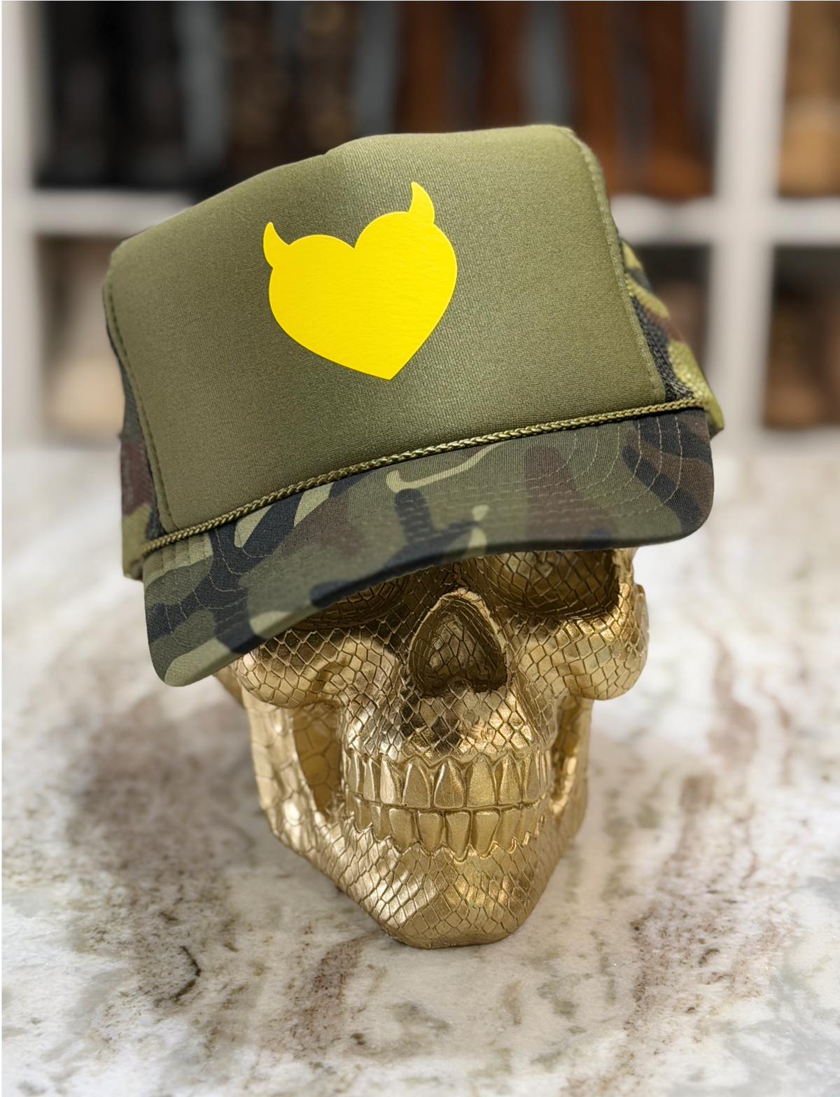 An olive green trucker hat with a yellow devil-heart logo on the front, displayed on a gold skull prop. The hat features a solid green front panel and a camouflage-patterned brim and side panels.