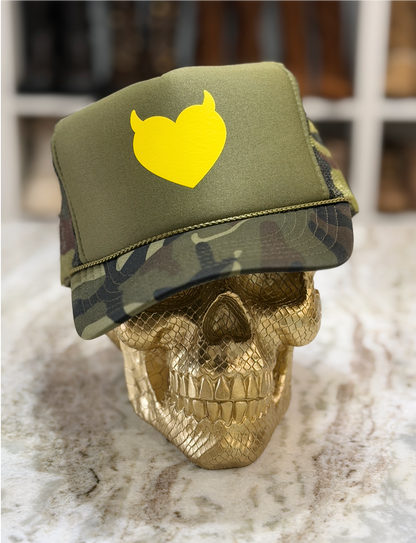 An olive green trucker hat with a yellow devil-heart logo on the front, displayed on a gold skull prop. The hat features a solid green front panel and a camouflage-patterned brim and side panels.