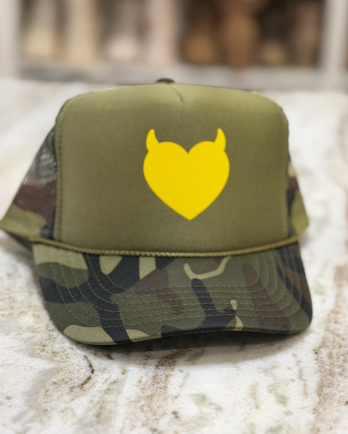 An olive green and camouflage trucker hat with a bright yellow devil-heart graphic on the front, resting on a light marble surface.