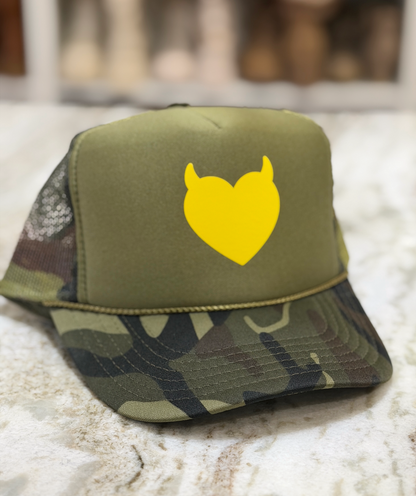 An olive green trucker hat with a yellow devil-heart graphic on the front, resting on a light marble surface.