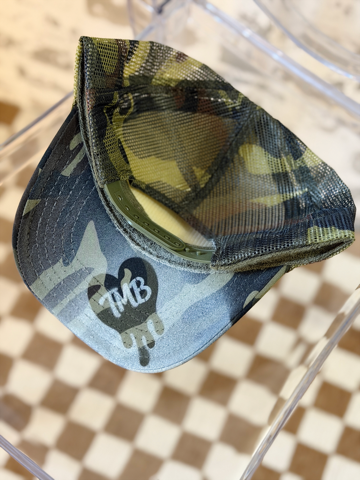 A camouflage trucker cap with a blue-gray and green camo pattern, featuring a black heart-shaped logo with 'TMB' in white on the brim. The cap is resting on a clear acrylic stand over a brown and beige checkered floor.
