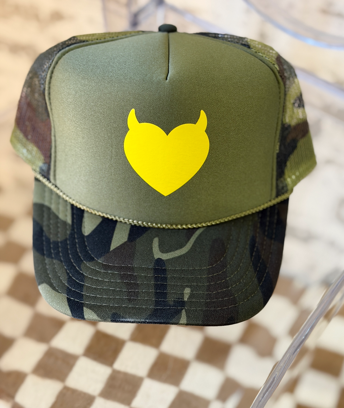 An olive green and camouflage trucker hat with a bright yellow devil-heart graphic on the front, displayed on a clear stand over a checkered floor.